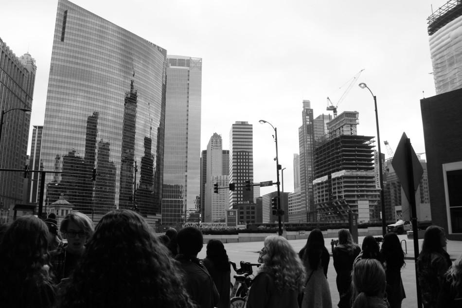 The buildings reflect off of the skyscraper on the overcast day. The students finished shopping and waited for their bus to arrive.