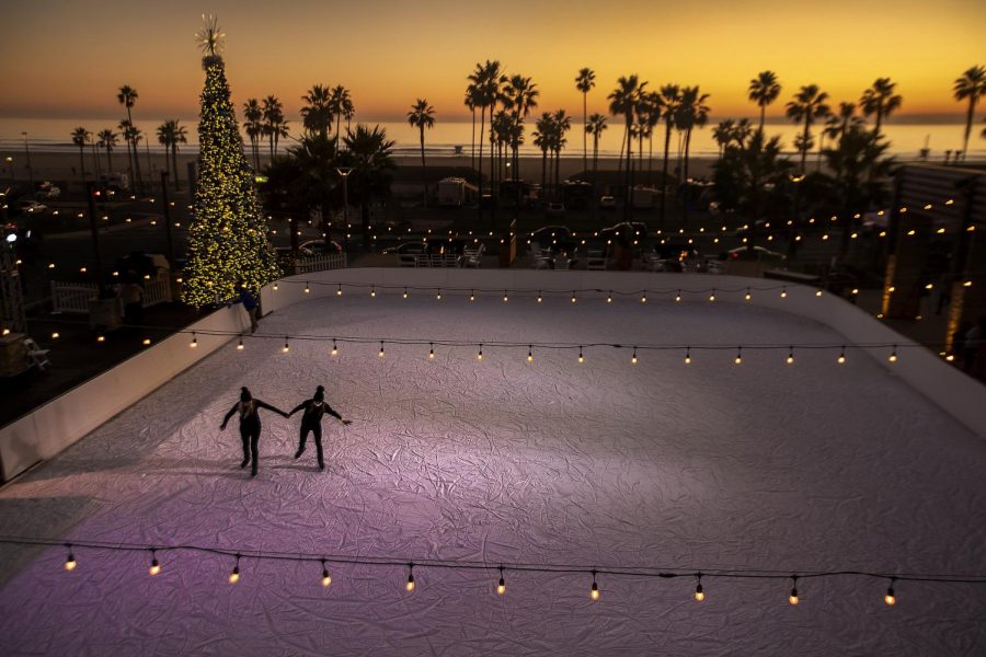 Two people hold hands while they ice skate outside under a christmas tree. The Christmas tree and fairy lights around the rink lighting up the way for the people.